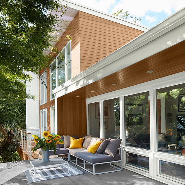A gray deck with a blue outdoor couch and yellow pillows, backs up to a one-story enclosed porch with brown wood siding and white painted trim, attached to a two-story home with light brown siding and white trim. A gray deck with a blue outdoor couch and yellow pillows, backs up to a one-story enclosed porch with brown wood siding and white painted trim, attached to a two-story home with light brown siding and white trim.