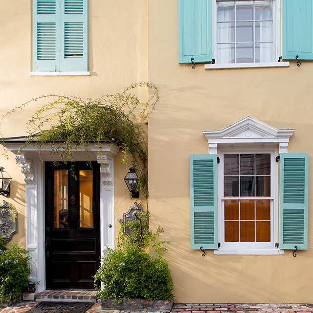 A pretty beige painted stucco home exterior with a soft peach undertone, blue-green shutters, white trim, and greenery around the black front door. A pretty beige painted stucco home exterior with a soft peach undertone, blue-green shutters, white trim, and greenery around the black front door.