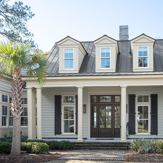 A beautiful gray-painted home features a white columned front porch and white trim, black shutters, a black glass-paned front door, dormer windows, and a palm tree in the front yard. A beautiful gray-painted home features a white columned front porch and white trim, black shutters, a black glass-paned front door, dormer windows, and a palm tree in the front yard.
