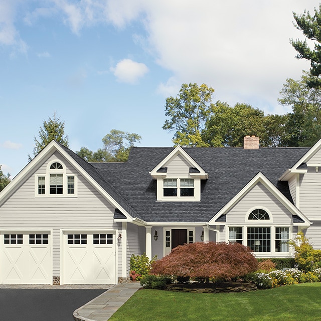 A pretty light gray-painted home exterior with white garage doors and trim, a lush green lawn and landscaping. A pretty light gray-painted home exterior with white garage doors and trim, a lush green lawn and landscaping.