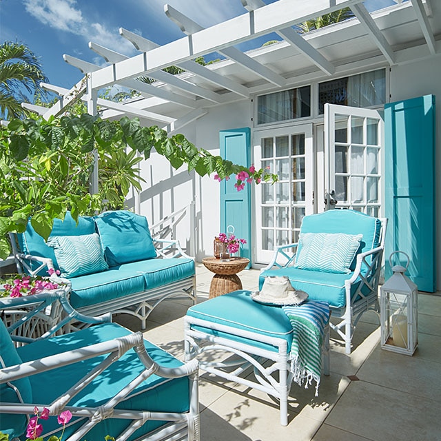 A pretty white-painted home exterior with turquoise shutters, an attached pergola and open french doors to a patio with turquoise cushioned furniture and tropical greenery. A pretty white-painted home exterior with turquoise shutters, an attached pergola and open french doors to a patio with turquoise cushioned furniture and tropical greenery.