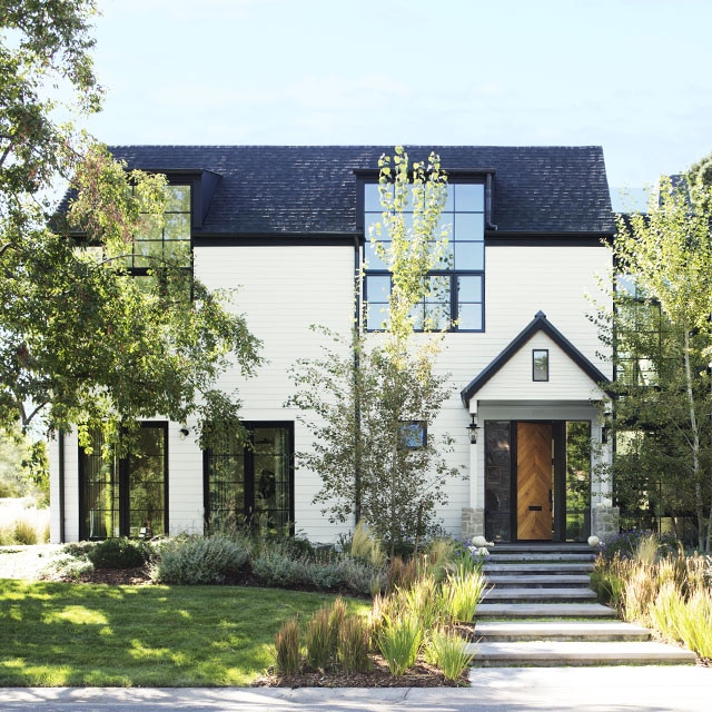 A beautiful home with white-painted siding on the left, stone exterior on the right side, and large black framed windows throughout, surrounded by greenery. A beautiful home with white-painted siding on the left, stone exterior on the right side, and large black framed windows throughout, surrounded by greenery.