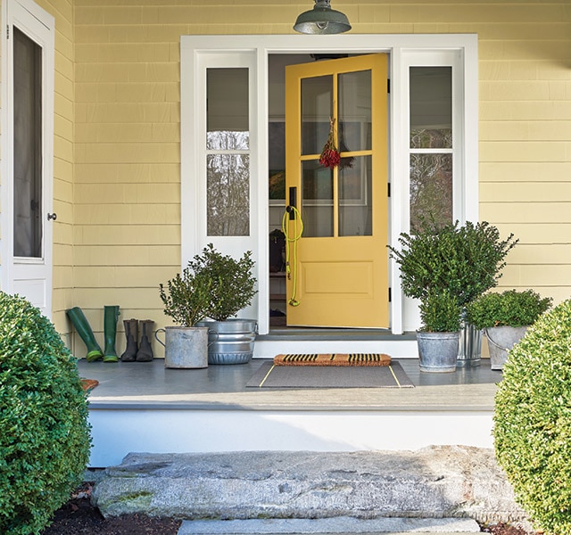 The front porch of a yellow-painted house with a yellow door, white trim, plants in metal pails, and two round shrubs out front.