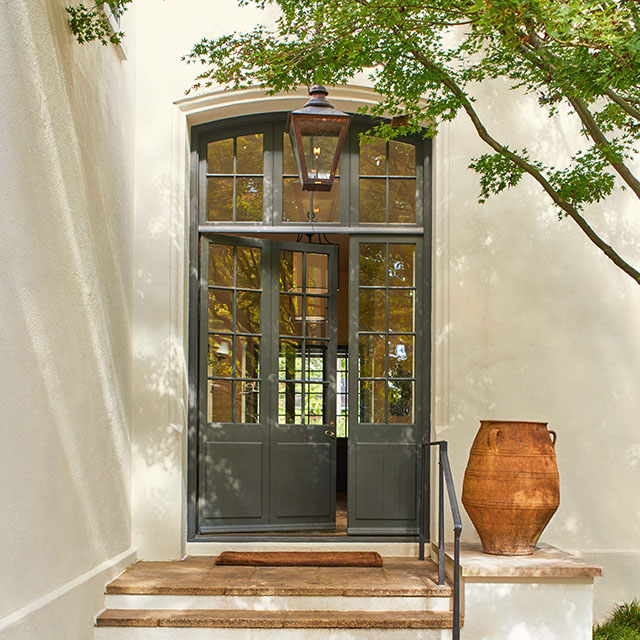 An off-white painted stucco house with an elegant dark green front door with glass panes and transom windows, ceramic tile steps and landing with a large clay pot, partially shaded by a tree. An off-white painted stucco house with an elegant dark green front door with glass panes and transom windows, ceramic tile steps and landing with a large clay pot, partially shaded by a tree.