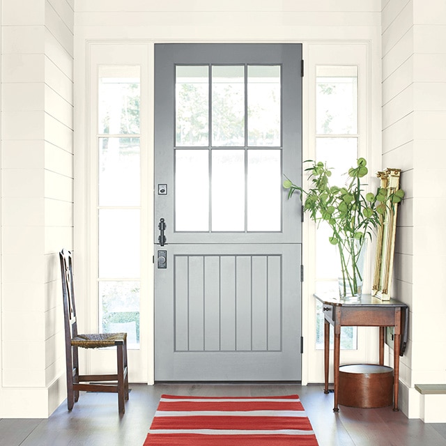 A bright and welcoming home entryway with a gray painted front door, white shiplap walls, a wood table and chair, and red-and-gray striped rug. A bright and welcoming home entryway with a gray painted front door, white shiplap walls, a wood table and chair, and red-and-gray striped rug.