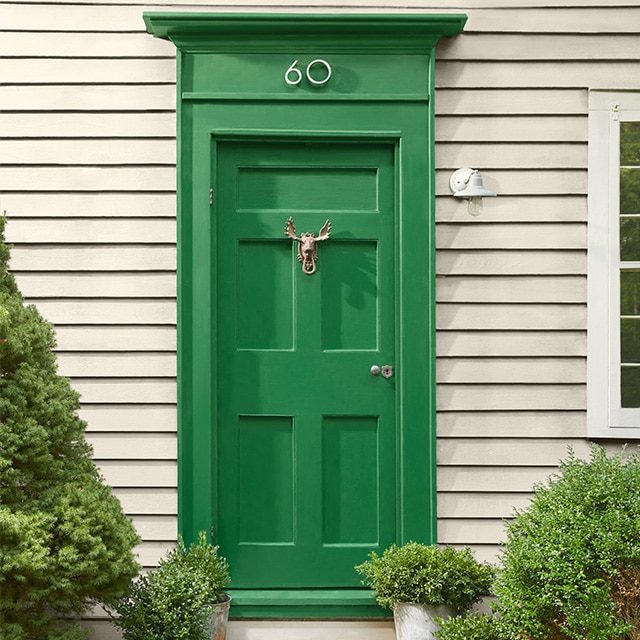 Warm greige-painted house siding, with a pretty green door flanked by shrubs, and white window trim. Warm greige-painted house siding, with a pretty green door flanked by shrubs, and white window trim.