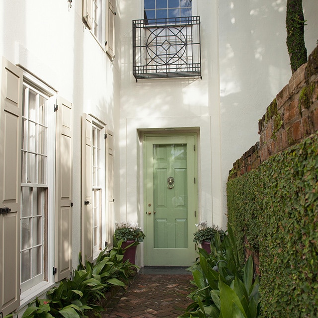 The front entrance of an elegant, white-painted house with beige shutters, a light-green front door, brick walkway and brick wall covered in foliage. The front entrance of an elegant, white-painted house with beige shutters, a light-green front door, brick walkway and brick wall covered in foliage.