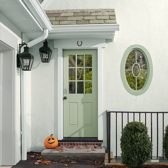 The side entrance of a white painted stucco home with a light-green painted door and oval-framed window, two black hanging lanterns, and a pumpkin on the doorstep. The side entrance of a white painted stucco home with a light-green painted door and oval-framed window, two black hanging lanterns, and a pumpkin on the doorstep.