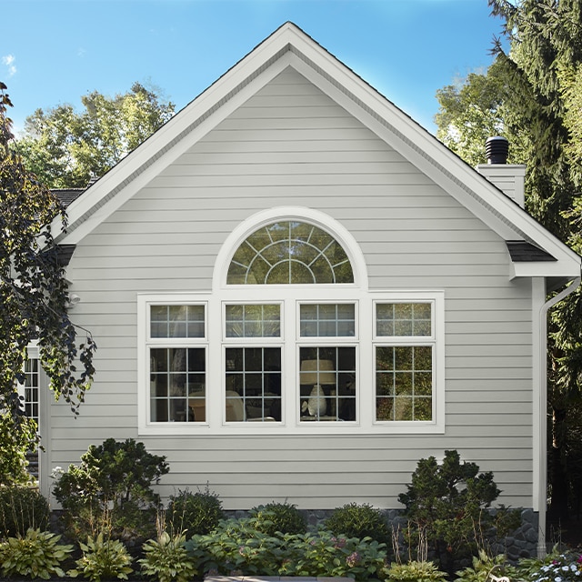 A house with light gray-painted exterior vinyl siding and white trimmed windows, surrounded by shrubs and trees. A house with light gray-painted exterior vinyl siding and white trimmed windows, surrounded by shrubs and trees.