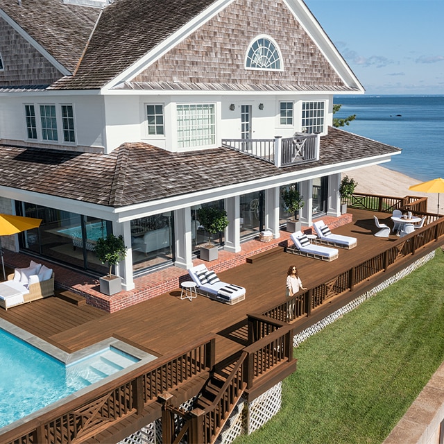 A lovely waterside home with white-painted siding and columns, taupe stained shingles, and a brown wrap around deck with lounge chairs, yellow umbrellas, and a pool. A lovely waterside home with white-painted siding and columns, taupe stained shingles, and a brown wrap around deck with lounge chairs, yellow umbrellas, and a pool.