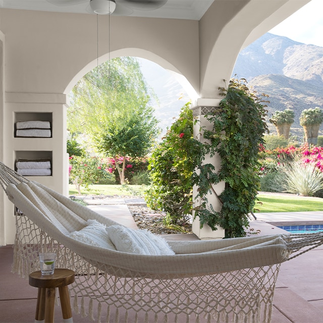 An inviting covered pool patio with white-painted stucco walls, climbing vines, and a hammock overlooking flowering red foliage and a scenic mountain range. An inviting covered pool patio with white-painted stucco walls, climbing vines, and a hammock overlooking flowering red foliage and a scenic mountain range.