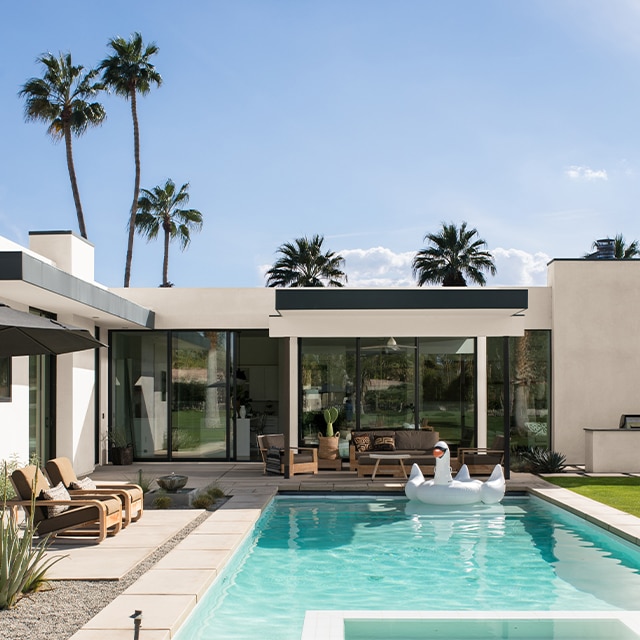 A modern, white-painted stucco house with black trim and palm trees features a beautiful backyard pool and patio. A modern, white-painted stucco house with black trim and palm trees features a beautiful backyard pool and patio.