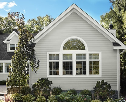 A house with light gray-painted exterior siding and white trimmed windows, surrounded by shrubs and trees. A house with light gray-painted exterior siding and white trimmed windows, surrounded by shrubs and trees.