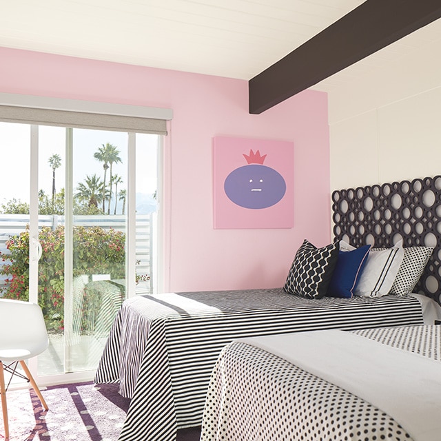 A bedroom with white-painted walls and ceiling, a cheery pink-painted accent wall, two twin beds with black and white polka dot and striped bedding, and a sliding door to a deck with views of palm trees and open sky. A bedroom with white-painted walls and ceiling, a cheery pink-painted accent wall, two twin beds with black and white polka dot and striped bedding, and a sliding door to a deck with views of palm trees and open sky.