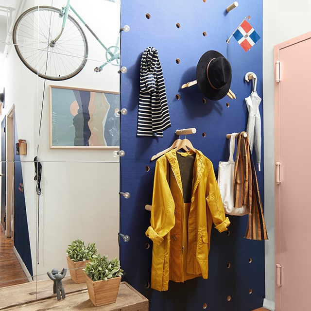 A loft entryway with a dark indigo blue pegboard wall holding hats, scarves, a jacket and umbrellas with a light pink front door. A loft entryway with a dark indigo blue pegboard wall holding hats, scarves, a jacket and umbrellas with a light pink front door.