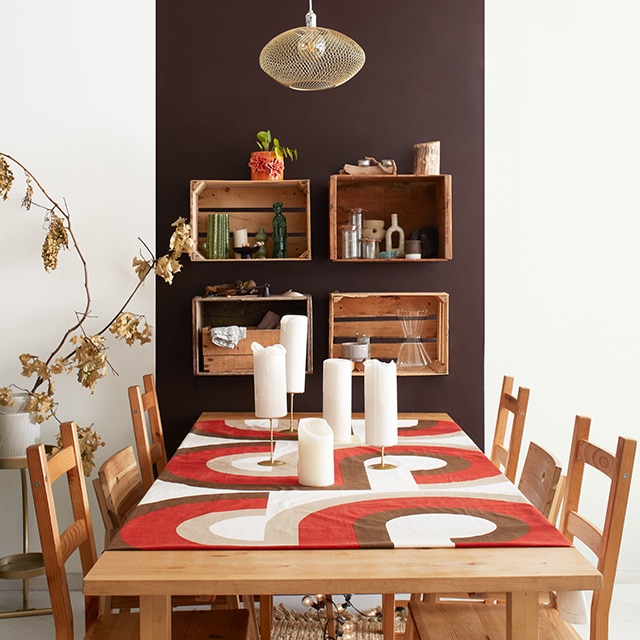 A modern apartment dining area with a dark brown-painted accent panel on a white wall, crate wall shelves, a wood table and chairs, and orange and brown table runner. A modern apartment dining area with a dark brown-painted accent panel on a white wall, crate wall shelves, a wood table and chairs, and orange and brown table runner.