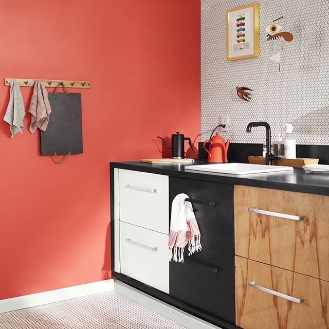 A sleek kitchen corner with a coral painted wall, a white tiled backsplash wall and floor, white upper wall and exposed pipes, and three sets of drawers: one painted white, the second black, and the third woodgrain. A sleek kitchen corner with a coral painted wall, a white tiled backsplash wall and floor, white upper wall and exposed pipes, and three sets of drawers: one painted white, the second black, and the third woodgrain.