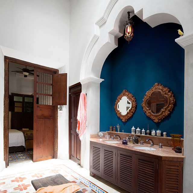 A pretty, white-painted bathroom with decorative molding, a double wooden vanity set in a blue-painted alcove, a colorful tile floor, wood bench and doors opening to a bedroom. A pretty, white-painted bathroom with decorative molding, a double wooden vanity set in a blue-painted alcove, a colorful tile floor, wood bench and doors opening to a bedroom.