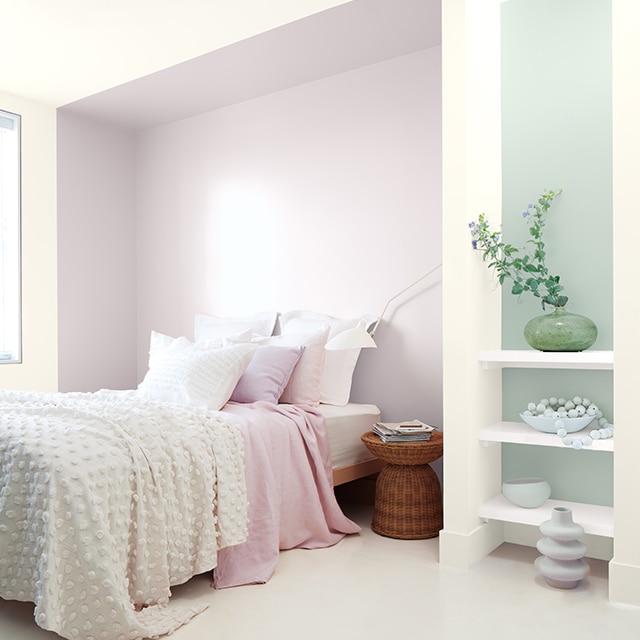 A white bedroom with a violet painted inset behind a bed with white and pink bedding, and a blue-green accent wall behind white shelving. A white bedroom with a violet painted inset behind a bed with white and pink bedding, and a blue-green accent wall behind white shelving.