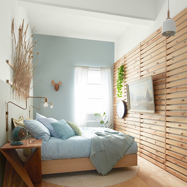 A cozy, sunlit bedroom with a light blue-painted accent wall, a two-tone muted silver and white wall behind a bed, a wood slatted wall with hanging artwork and a raised wood floor. A cozy, sunlit bedroom with a light blue-painted accent wall, a two-tone muted silver and white wall behind a bed, a wood slatted wall with hanging artwork and a raised wood floor.