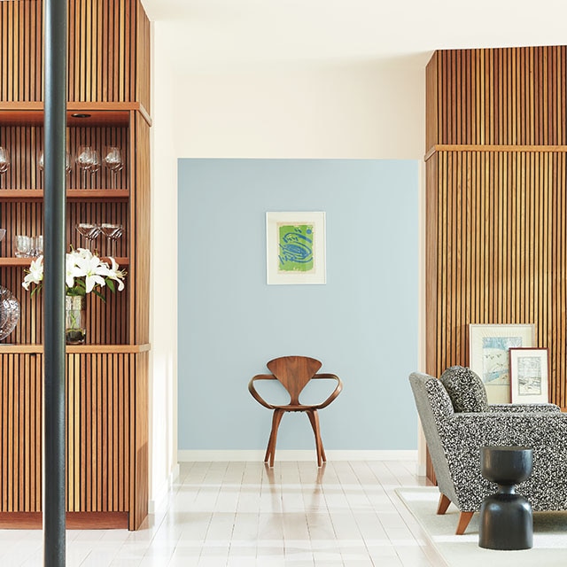 A bright, open living space with wood grooved paneling and cabinets, white walls, a black chair, and a modern wood chair against a light blue-painted hallway wall. A bright, open living space with wood grooved paneling and cabinets, white walls, a black chair, and a modern wood chair against a light blue-painted hallway wall.