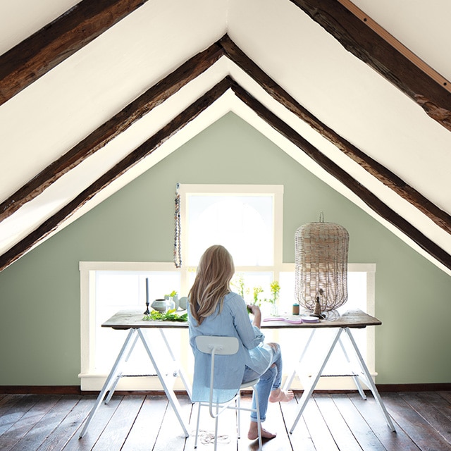 A woman sits at a contemporary desk in a small office space with a sloped white ceiling with wooden beams and a soft green-painted accent wall framing windows. A woman sits at a contemporary desk in a small office space with a sloped white ceiling with wooden beams and a soft green-painted accent wall framing windows.