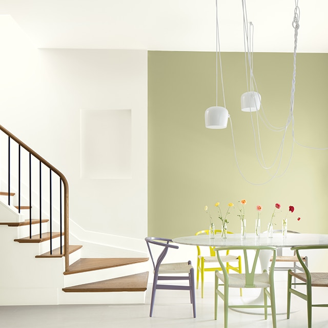 An open dining area with a light-green painted accent wall, white walls and ceiling, multi-colored chairs and a white table near a white open staircase with wood steps and banister. An open dining area with a light-green painted accent wall, white walls and ceiling, multi-colored chairs and a white table near a white open staircase with wood steps and banister.
