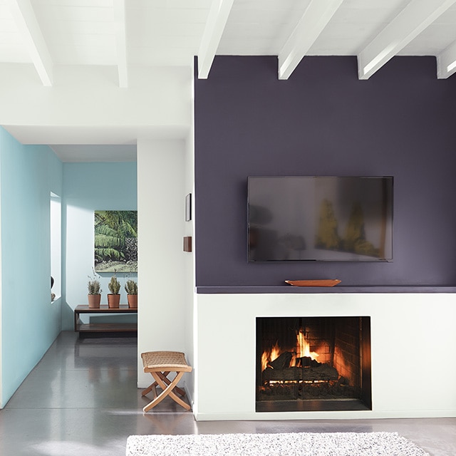 A modern white living room space with a rich, purple-painted accent wall and mantel over an active fireplace, and pretty blue hallway and back walls. A modern white living room space with a rich, purple-painted accent wall and mantel over an active fireplace, and pretty blue hallway and back walls.