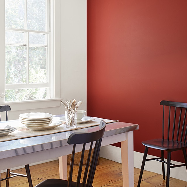 A dining room with a red-painted accent wall, a white wall, trim and ceiling, wood floors, a white table set with white dishes, and black spindle-back chairs. A dining room with a red-painted accent wall, a white wall, trim and ceiling, wood floors, a white table set with white dishes, and black spindle-back chairs.