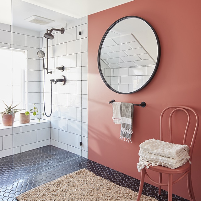 A bathroom with a red-painted accent wall, round mirror with black frame, a red-painted chair holding towels, white tiled walls and black tiled flooring with a beige bathmat. A bathroom with a red-painted accent wall, round mirror with black frame, a red-painted chair holding towels, white tiled walls and black tiled flooring with a beige bathmat.
