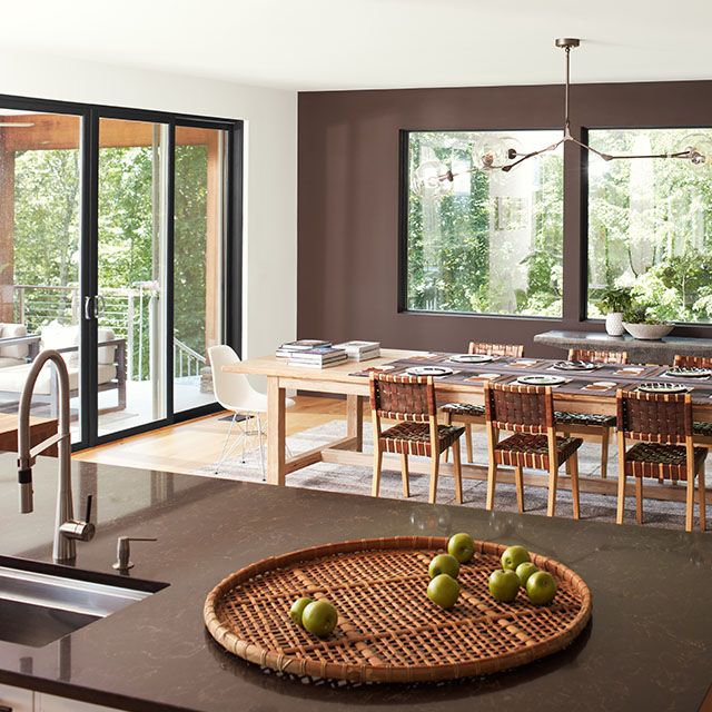 An open, modern dining room with a brown-painted accent wall and three large windows, a white ceiling and side wall with sliding glass doors, and brown granite kitchen island in the forefront. An open, modern dining room with a brown-painted accent wall and three large windows, a white ceiling and side wall with sliding glass doors, and brown granite kitchen island in the forefront.