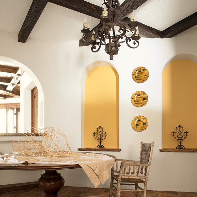 An airy, white-painted dining room with two yellow-painted alcoves, yellow decorative plates, dark wood ceiling beams, a rustic metal chandelier, and a round wood table and chair. An airy, white-painted dining room with two yellow-painted alcoves, yellow decorative plates, dark wood ceiling beams, a rustic metal chandelier, and a round wood table and chair.