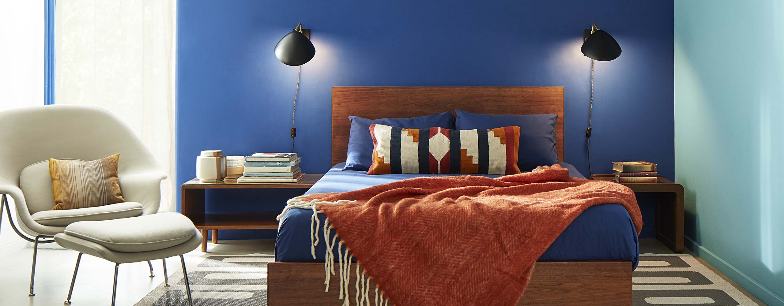A bedroom with two blue-painted accent walls, a wooden bed frame, rust colored throw, contemporary beige chair, and reading lamps on both sides of bed. A bedroom with two blue-painted accent walls, a wooden bed frame, rust colored throw, contemporary beige chair, and reading lamps on both sides of bed.