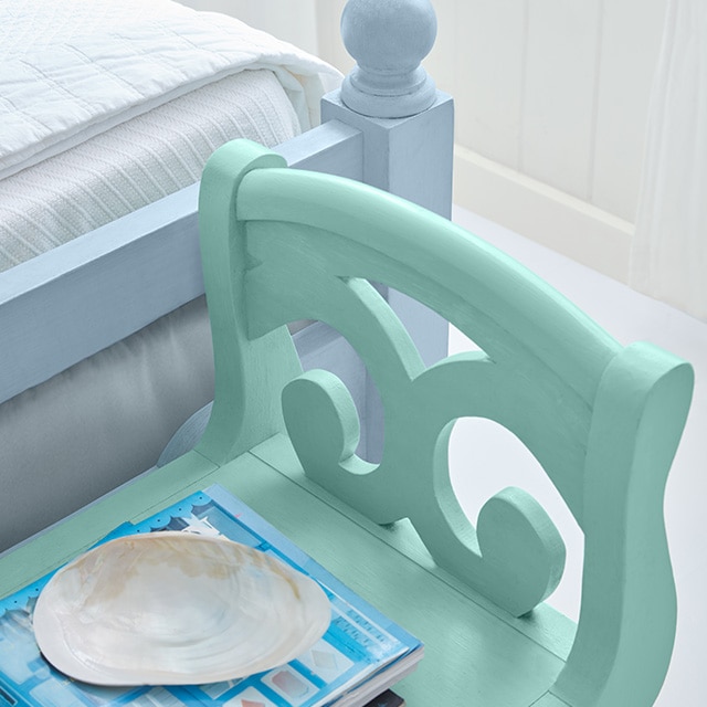 The corner of a sky blue-painted wood bed frame and a light turquoise bench near a white shiplap wall. The corner of a sky blue-painted wood bed frame and a light turquoise bench near a white shiplap wall.