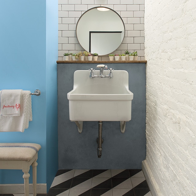 This eclectic bathroom features a blue painted wall, a white brick wall, a white sink against a gray wall with white tile and a mirror above, and a black-and-white chevron floor. This eclectic bathroom features a blue painted wall, a white brick wall, a white sink against a gray wall with white tile and a mirror above, and a black-and-white chevron floor.