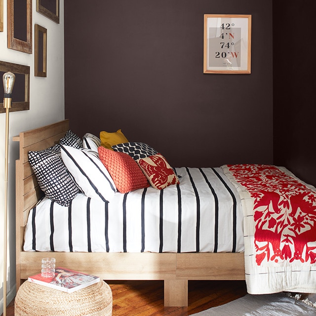 A cozy bedroom with deep chocolate-hued walls, a white-painted gallery wall, a bed with black and white striped bedding, pillows and red printed blanket, and a wicker ottoman. A cozy bedroom with deep chocolate-hued walls, a white-painted gallery wall, a bed with black and white striped bedding, pillows and red printed blanket, and a wicker ottoman.