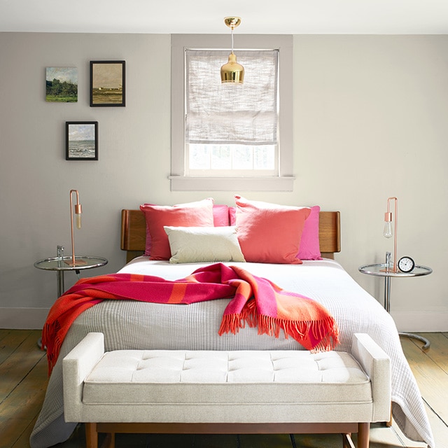 Bright coral pillows and a throw really pop in this serene, greige painted bedroom with an off-white quilt on the bed, an off-white upholstered bench, and natural wood floor. Bright coral pillows and a throw really pop in this serene, greige painted bedroom with an off-white quilt on the bed, an off-white upholstered bench, and natural wood floor.