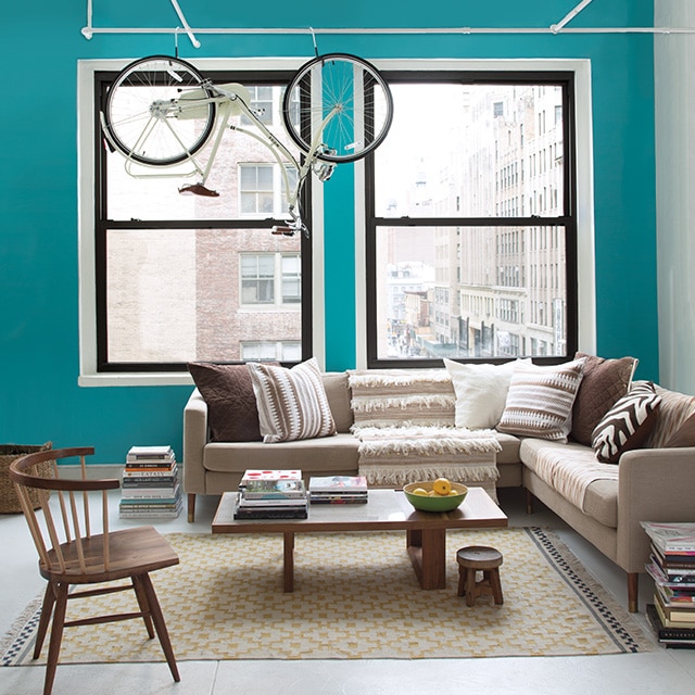 A bright, city apartment living room with a turquoise-painted accent wall, white side wall and trim, two large windows, beige and brown furnishings, and a bike hanging from exposed pipes. A bright, city apartment living room with a turquoise-painted accent wall, white side wall and trim, two large windows, beige and brown furnishings, and a bike hanging from exposed pipes.
