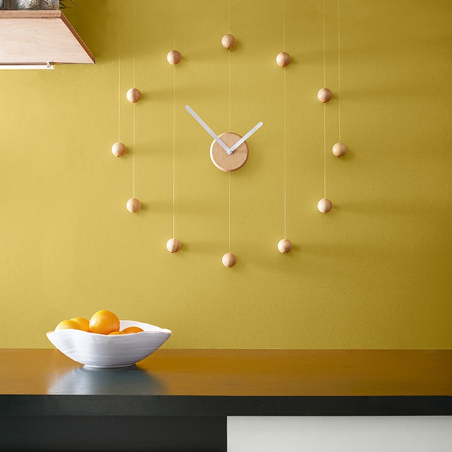A yellow ochre painted kitchen wall features a minimalist wood clock over a brown countertop, with a stainless-steel oven and white drawer underneath. A yellow ochre painted kitchen wall features a minimalist wood clock over a brown countertop, with a stainless-steel oven and white drawer underneath.