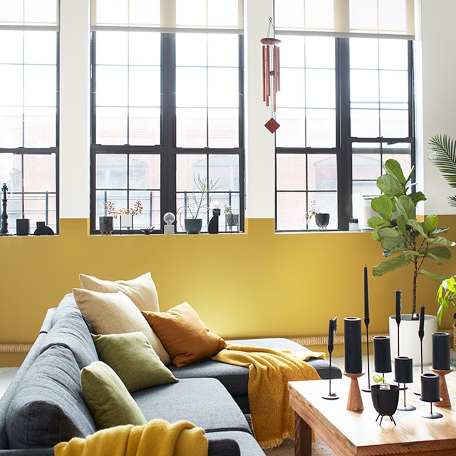 An airy, loft-style living room with a white and yellow accent wall with tall windows, a white ceiling with exposed pipes, and gray, yellow, and wood modern furnishings. An airy, loft-style living room with a white and yellow accent wall with tall windows, a white ceiling with exposed pipes, and gray, yellow, and wood modern furnishings.