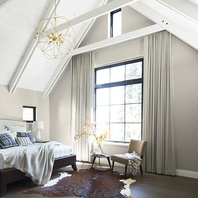 A sunlit bedroom with light gray-painted walls, a white cathedral ceiling with beams and a large paned window. A sunlit bedroom with light gray-painted walls, a white cathedral ceiling with beams and a large paned window.