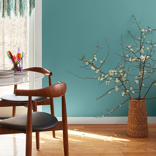 A pretty, teal-painted dining room wall with white trim, a teal macrame doorway hanging, flowered branches in a basket, modern furniture, and a hardwood floor. A pretty, teal-painted dining room wall with white trim, a teal macrame doorway hanging, flowered branches in a basket, modern furniture, and a hardwood floor.
