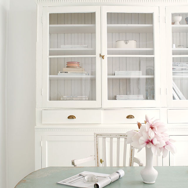 A shabby chic style dining room with white-painted walls, a white cabinet and chair, a light-blue distressed table with a vase of flowers, and light-gray painted wood floor. 