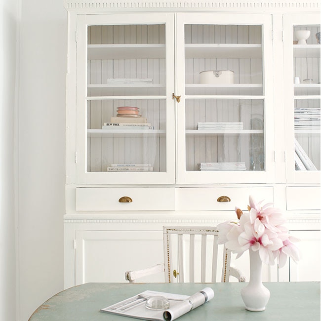 A shabby chic style dining room with white-painted walls, a white cabinet and chair, a light-blue distressed table with a vase of flowers, and light-gray painted wood floor. 