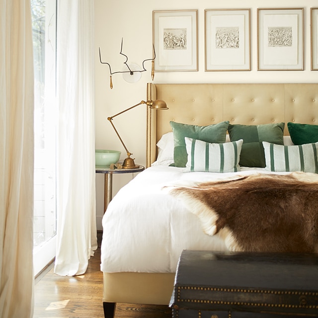 An off-white-painted bedroom with white and green bedding and pillows, a tan upholstered headboard and off-white drapes. An off-white-painted bedroom with white and green bedding and pillows, a tan upholstered headboard and off-white drapes.