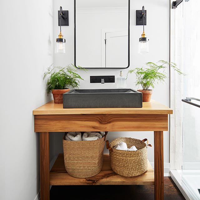 A corner of a white-painted bathroom with a wood vanity, a black square sink, modern black lights and mirror, and baskets on lower shelf. A corner of a white-painted bathroom with a wood vanity, a black square sink, modern black lights and mirror, and baskets on lower shelf.