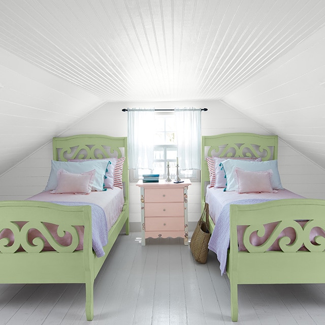 A cozy white-painted attic bedroom with shiplap walls and ceiling shows off two green twin beds with multi-colored pastel bedding, flanking a window and small pink chest of drawers. A cozy white-painted attic bedroom with shiplap walls and ceiling shows off two green twin beds with multi-colored pastel bedding, flanking a window and small pink chest of drawers.