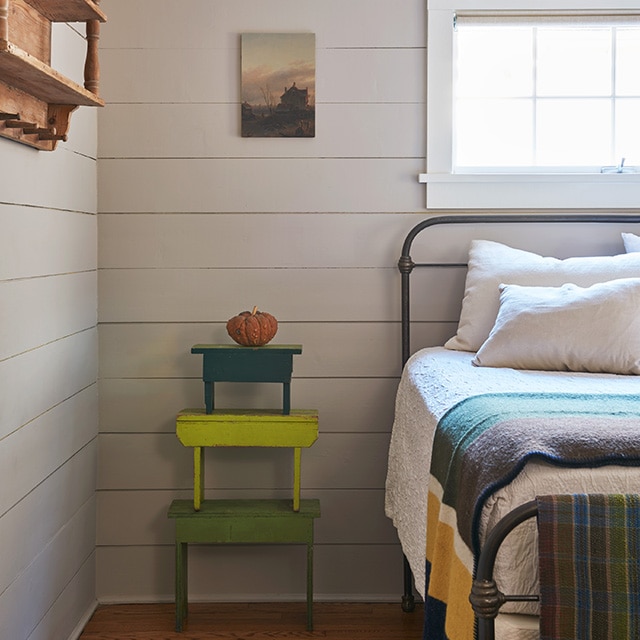 A comfy bedroom corner with beige-painted shiplap walls, a black-framed iron bed with multi-colored blankets under a white trimmed window and colorful stacked benches. A comfy bedroom corner with beige-painted shiplap walls, a black-framed iron bed with multi-colored blankets under a white trimmed window and colorful stacked benches.