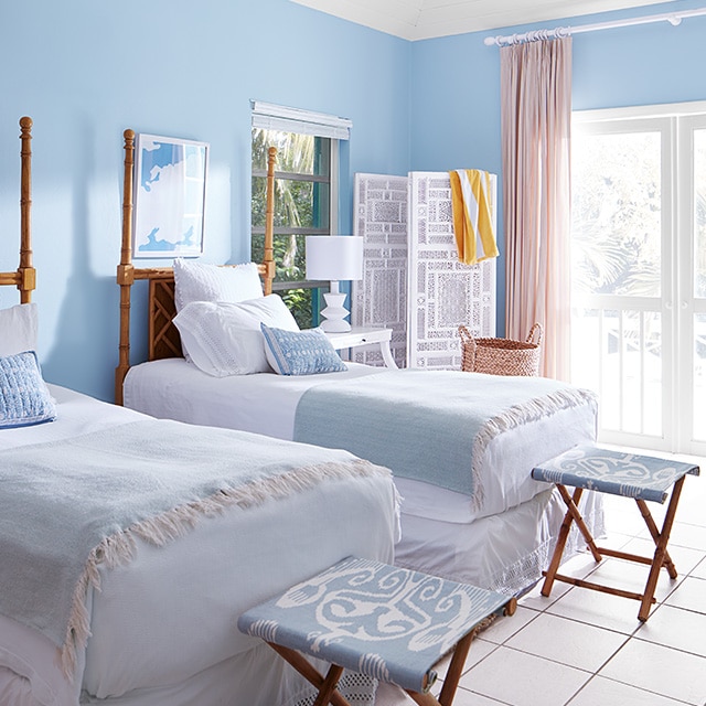 A bedroom with blue painted walls, a white ceiling and trim, twin beds and folding stools in blue and white decor. A bedroom with blue painted walls, a white ceiling and trim, twin beds and folding stools in blue and white decor.