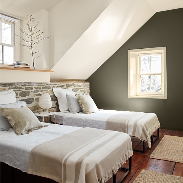 A cozy white-painted bedroom with a moss green accent wall, two twin beds in front of a low stone wall, hardwood floors and two small windows. A cozy white-painted bedroom with a moss green accent wall, two twin beds in front of a low stone wall, hardwood floors and two small windows.
